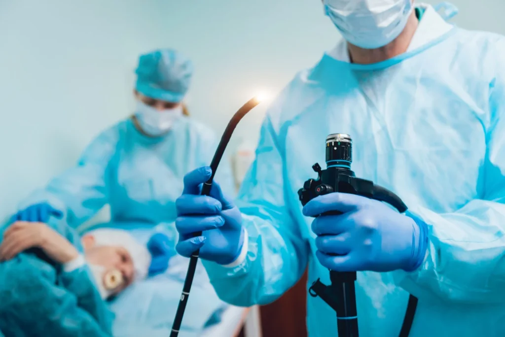 A doctor is holding an endoscope. In the background, a nurse is tending to a patient.